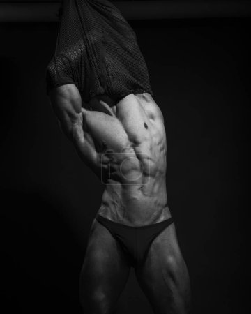 Foto de Hombre musculoso sexy en traje de baño sobre fondo negro. Retrato en blanco y negro del modelo masculino quitándose la camisa. Torso masculino en estudio. - Imagen libre de derechos