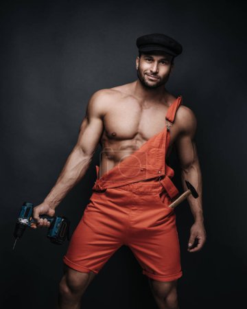 Handsome man in red jumpsuit and black beret on dark background in studio. Sexy handyman with tools. Male worker portrait with hammer and drill.