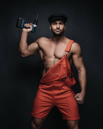 Handsome man in red jumpsuit and black beret on dark background in studio. Sexy handyman with tools. Male worker portrait with screwdriver.