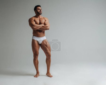 Sexy muscular man in white underwear posing in studio. Handsome bodybuilder standing on grey background. Big male model showing his muscles.