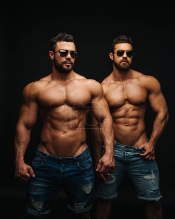 Foto de Dos tíos sin camisa con fondo negro. Modelos de fitness pareja en jeans y gafas de sol. Dos hombres guapos musculosos con seis abdominales en el estudio. - Imagen libre de derechos