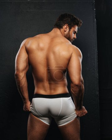 Hombre musculoso desnudo en ropa interior de boxeador blanco sobre fondo negro. Sexy espalda de culturista en el estudio. Parte posterior del modelo masculino muscular sosteniendo su ropa interior.