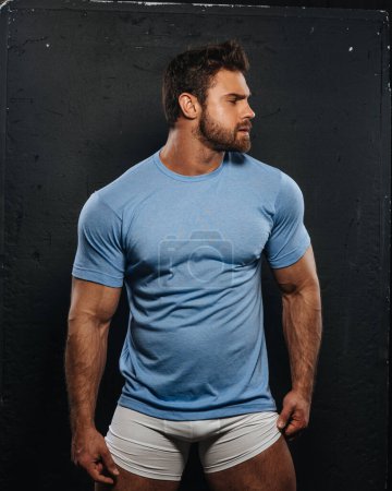 Téléchargez les photos : Bearded muscled man in blue shirt and white boxers looking to right side. Sexy fitness guy wearing casual clothes at black background. Attractive bodybuilder in studio. - en image libre de droit