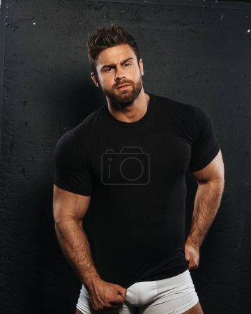 Photo for Bearded handsome man in black shirt and white boxers at black background. Sexy muscled guy in casual underwear in studio. Male fitness model posing in relaxing outfit. - Royalty Free Image