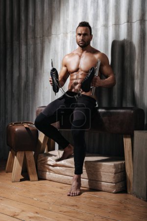 Sexy shirtless man posing on gymnastics pommel horse with ice skates in his hands. Muscular male model in black leggings in the studio. Nude handsome guy with sports equipment.
