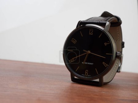 Foto de Water resistant black watch with leather strap placed on right side a desk with white wall - Imagen libre de derechos