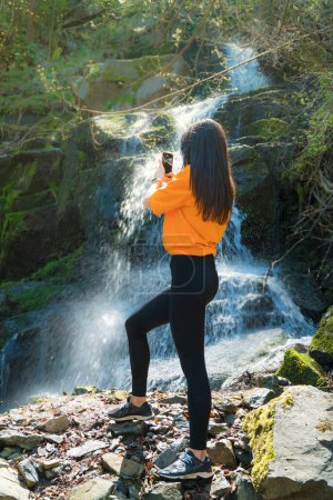 Foto de Mujer de pie frente a la cascada y tomando fotos con el teléfono - Imagen libre de derechos