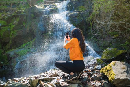 Foto de Mujer de pie frente a la cascada y tomando fotos con el teléfono - Imagen libre de derechos