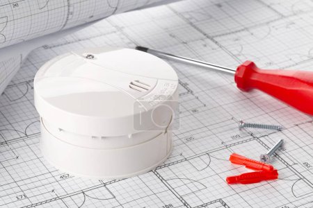 Smoke detector or fire alarm sensor on white architectural plans background with tools and screws, house safety or security concept