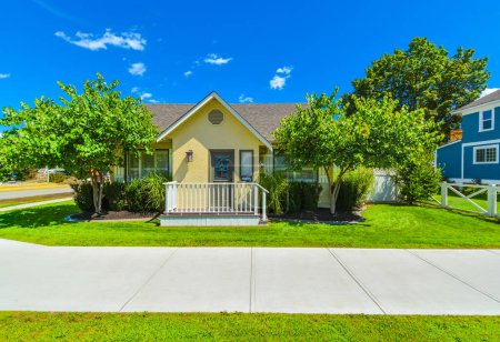 Façade de la petite maison familiale par une journée ensoleillée avec des marches de porte en face. Maison jaune avec petite cour et sentier en béton sur fond de ciel bleu.