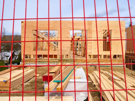 Wooden frame and walls of new house behind the red fence grid.