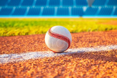 Photo for A single white baseball with red stitching lies on a white field line painted on orange dirt in an empty stadium during daylight. - Royalty Free Image