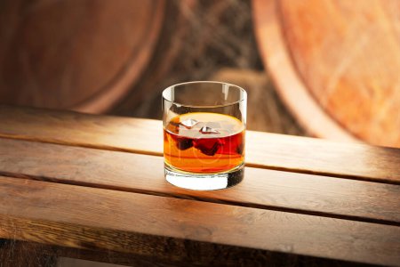 Photo for Glass of fine aged whiskey with ice in liquid. Old, dusty distillery cellar with many wooden barrels covered by dense cobwebs. Maturation of finest alcohol beverage takes time. Seasoning hard liquor. - Royalty Free Image