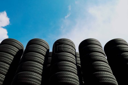 Photo for Many stacks of car tires, both new or used, are piled high against a bright blue sky. Black rubber tires. Concept of auto service, wheels changing, tire store. Tires for sale. Transportation. - Royalty Free Image