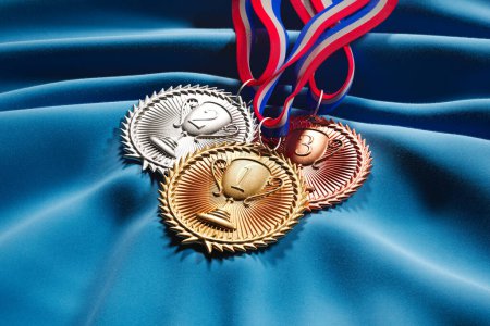 Foto de Colección de medallas de oro, plata y bronce con cintas de colores que descansan sobre un fondo de tela azul suave. Perfecto para premios, reconocimiento o conceptos de competición. - Imagen libre de derechos