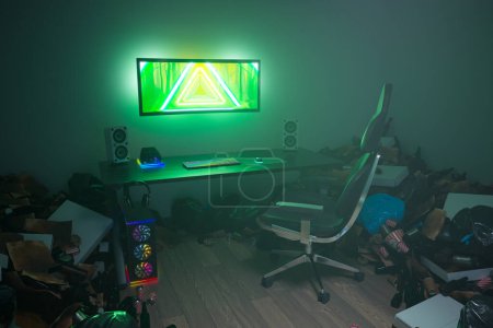 Foto de Imagen deprimente con una sala de juegos desordenada. Potente set de ordenador para jugadores con un gran monitor y cómodo sillón. La habitación está llena de basura como cajas de pizza y botellas. Pereza - Imagen libre de derechos