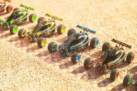 Mehrere farbenfrohe ferngesteuerte Buggy-Autos, die hintereinander an der Startlinie in der Wüste auf das Rennen warten. Spielzeugkreislauf beginnt. Heißer, sonniger Tag und trockene Sandrennbahn. Ansicht von oben.
