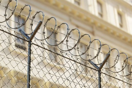 Une image montre une clôture avec du fil de fer barbelé qui entoure un important bâtiment stratégique. Il peut s'agir d'une prison, d'un objet militaire ou d'une ambassade, ce qui donne un sentiment de confinement ou de protection.
