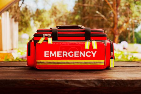 Photo for Professional red emergency first aid bag on a wooden table outdoors. Doctor's kit stacked with equipment and ready for fast response in a green, lush, natural environment. Saving lives. - Royalty Free Image