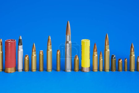 A huge row of different bullet types standing on the concrete floor. Huge variety of ammunition calibers. Handgun, carabine, automatic, shotgun, and more. Deadly combat force. Blue background