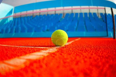 Téléchargez les photos : Vue rapprochée de dessous d'une balle de tennis jaune couchée sur un court orange avec un filet en arrière-plan et des tribunes bleues. L'image est éclairée par la lumière naturelle du jour. - en image libre de droit
