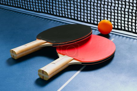 Photo for A detailed view of two table tennis paddles resting on a blue table, with an orange ball nearby, ready for a game. The net adds an element of competition and excitement to the scene. - Royalty Free Image