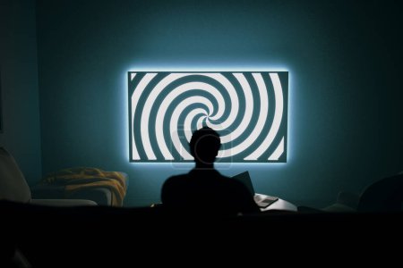 A 3D rendering of a dark room with a silhouette of a person sitting and watching TV. The TV screen displays a hypnotizing spiral. This image represents manipulation and the power of media.