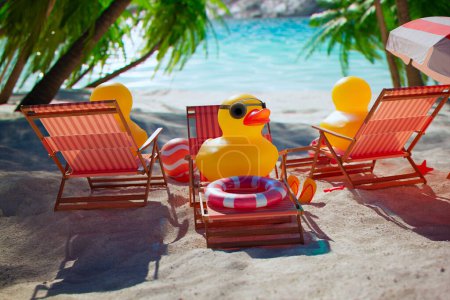 Téléchargez les photos : Canard en caoutchouc dans des lunettes de soleil sur la plage. Jolis jouets jaunes sur une serviette de plage et chaises longues sous les parasols. Palmiers et une mer turquoise calme en arrière-plan. Ambiance joyeuse. Détendez-vous. Vacances - en image libre de droit