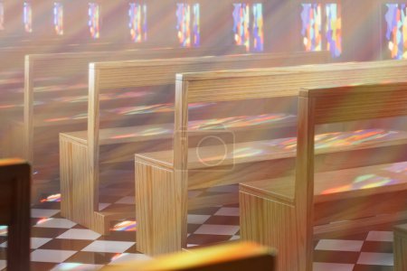 An empty chapel with rows of wooden benches bathed in warm sunlight streaming through stained glass windows. The vibrant god rays create a sense of religious and spiritual significance