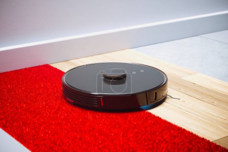 Photo for This efficient robotic vacuum cleaner seamlessly navigates from a plush red carpet to sleek hardwood flooring, illustrating its versatility and high-tech design for modern home upkeep. - Royalty Free Image