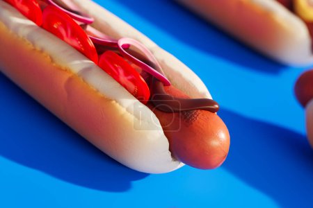 Photo for A close-up shot of a tasty hot dog, topped with onions, tomatoes and BBQ sauce. The hot dog is presented on a striking blue background with other hot dogs subtly blurred in the background. - Royalty Free Image