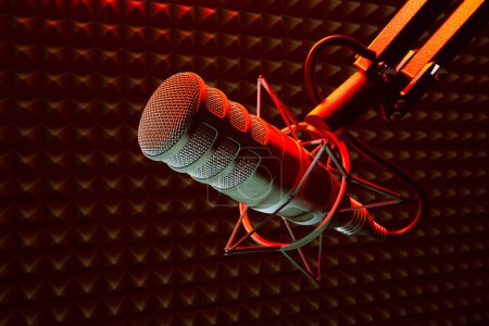 Vista de cerca que captura los detalles de un micrófono de estudio profesional en un soporte de choque rojo, frente a la espuma absorbente de sonido de un estudio de grabación bien iluminado.