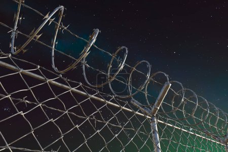 Photo for Dramatic close-up of barbed wire crowning a fence, starkly outlined by the celestial backdrop of a star-filled sky, embodying security and confinement. - Royalty Free Image