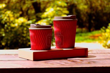 Foto de Vista de cerca que muestra un dúo de tazas de café de color rojo brillante con tapas, sobre una mesa de madera rústica, en medio de un fondo verde natural suavemente borroso. - Imagen libre de derechos