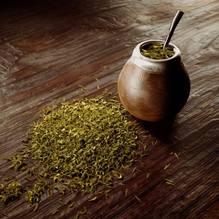 Close-up view of a traditional wooden gourd filled with Yerba Mate, accompanied by a metal bombilla; a loose-leaf herb spread on the rustic wooden background enhances the scene.