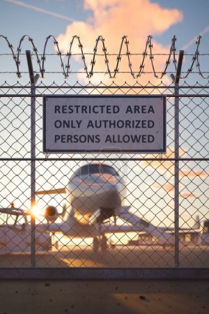 Téléchargez les photos : Silhouette d'une clôture de sécurité barbelée surmontée d'un fil de fer barbelé et d'un panneau de zone réglementée dans un aéroport alors qu'un avion est vu contre un ciel couchant vif. - en image libre de droit