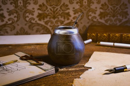 A meticulously arranged scene featuring an old-fashioned yerba mate tea set with gourd and bombilla beside a pen-laden sketchbook, all set against a retro-styled wallpaper backdrop.