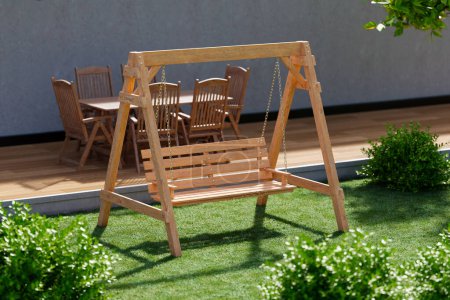 Photo for Captivating backyard escape featuring a wooden swing bench bathed in sunlight, complemented by a lush lawn and cozy patio arrangement with chairs and a table. - Royalty Free Image