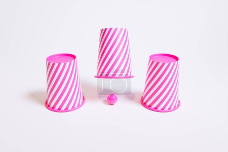 A dynamic arrangement of three pink and white striped cups, one inverted, alongside a matching small ball, all set against a pristine white backdrop, showcasing minimalism and vibrancy.