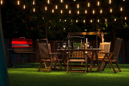 Bezaubernde Hinterhof-Szene am Abend mit einem stilvollen Outdoor-Esszimmer mit eleganten Holzmöbeln, stimmungsvoller Lichterkette und einem einladenden Grill.