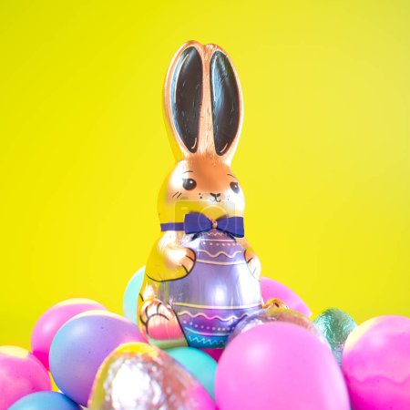 Foto de Pieza central de conejo de Pascua metálica exquisitamente brillante rodeada por una variedad de huevos vibrantes y multicolores colocados artísticamente sobre un fondo amarillo soleado. - Imagen libre de derechos