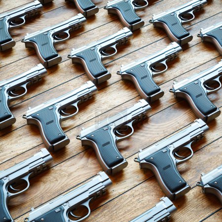 Photo for This photo captures an array of sleek silver semi-automatic handguns meticulously lined up on a wooden table, showcasing a striking contrast between the cold metal and the warm wood textures. - Royalty Free Image