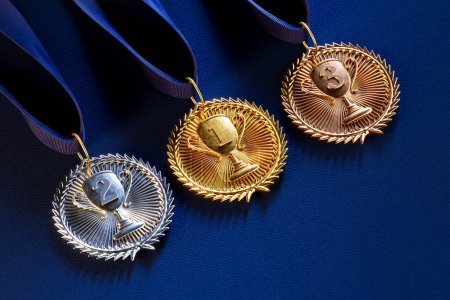 Foto de Primer plano de medallas campeonas de oro, plata y bronce con cintas sobre un lujoso fondo de tela azul profundo, personificando el pináculo del éxito competitivo. - Imagen libre de derechos