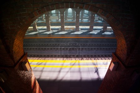 Una composición evocadora resalta las texturas y formas contrastantes a medida que la luz del sol fluye a través de una ventana arqueada, ofreciendo una vista voyeurística de una plataforma ferroviaria ocupada debajo llena de viajeros..
