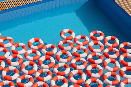 Luftaufnahme, die einen auffallenden Kontrast mehrerer roter und weißer Rettungsringe einfängt, die geordnet über das ruhige blaue Wasser eines Swimmingpools verteilt sind und Sicherheit und Freizeit symbolisieren.