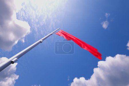 Téléchargez les photos : Un remarquable drapeau rouge ondule dans le vent au sommet d'un poteau métallique robuste, vif sur fond azur avec des nuages tortueux, véhiculant un message de prudence et de vigilance. - en image libre de droit