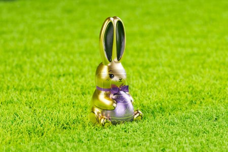 Foto de Imagen cautivadora de un reluciente conejito de Pascua dorado adornado con una cinta púrpura real, encaramado en la cima de una exuberante hierba verde artificial, encarnando el espíritu de las celebraciones de primavera. - Imagen libre de derechos