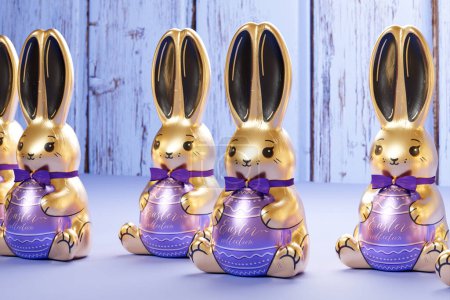Foto de Exquisita línea de chocolates de conejo de Pascua bañados en oro envueltos en tonos púrpuras, que simbolizan regalos festivos de lujo, posados en una superficie de madera rústica para las vacaciones de primavera. - Imagen libre de derechos
