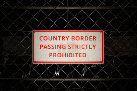 Intensive Nahaufnahme eines krassen Warnschildes auf einem sicheren Maschendrahtzaun, der eine nationale Grenze markiert, symbolisiert strenge Vorschriften und ein erzwungenes Verbot des unbefugten Grenzübertritts.