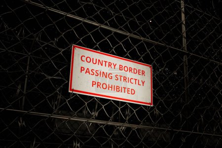 Photo for A vivid red and white prohibition sign declares COUNTRY BORDER PASSING STRICTLY PROHIBITED on a chain-link fence, highlighting the stark contrast against the dark twilight backdrop. - Royalty Free Image